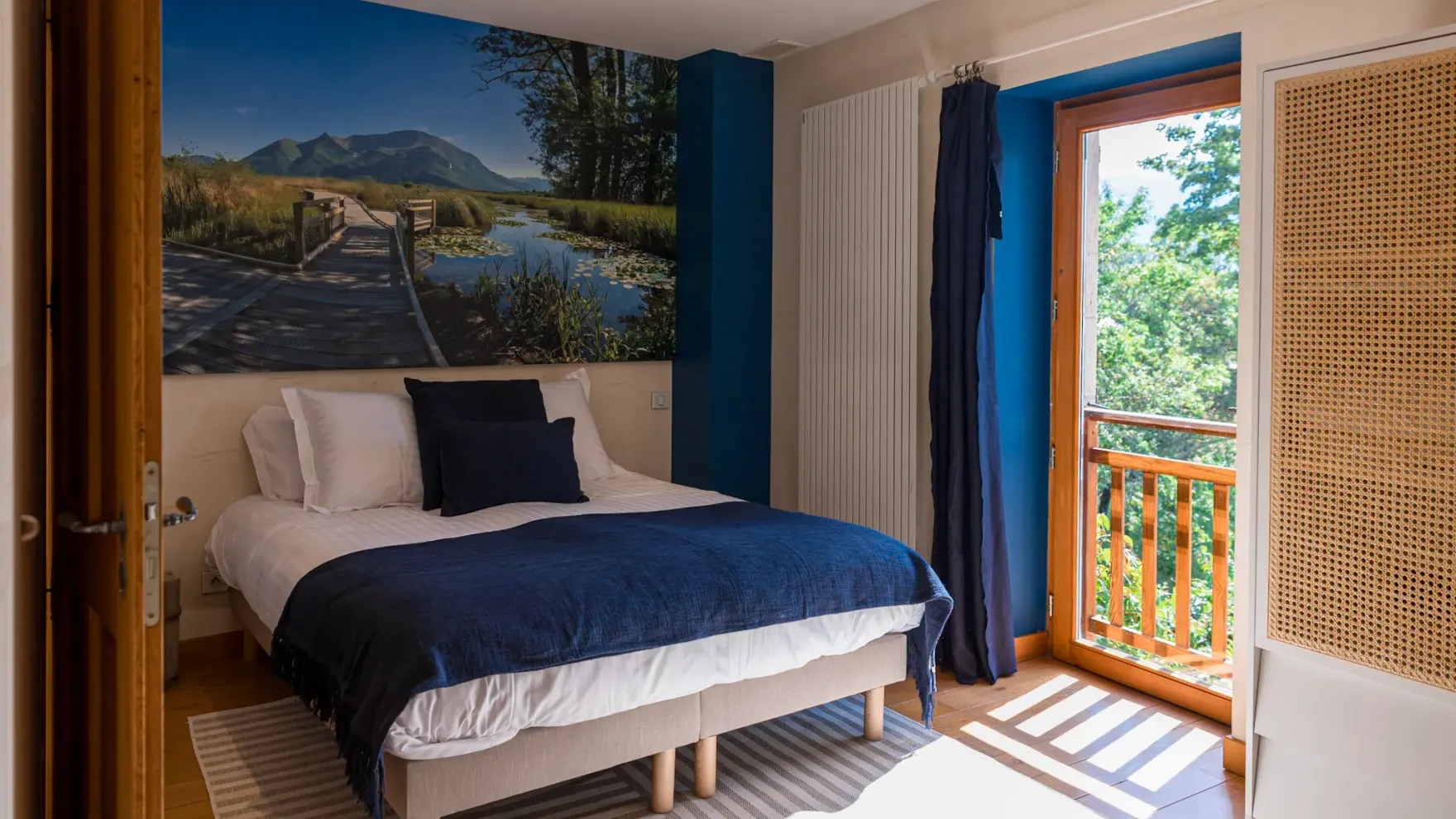 Chambre avec un lit double recouvert d'une couverture bleue et de coussins, un mur décoré d'une grande photo de paysage naturel, une porte-fenêtre ouverte sur un balcon en bois avec vue sur des arbres